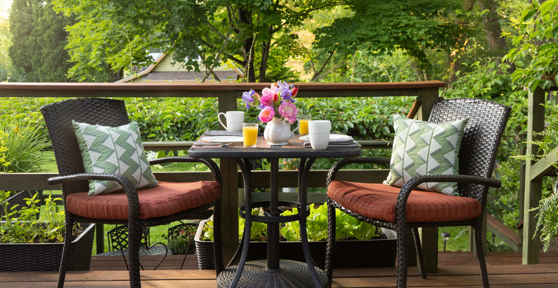 An outdoor dining table and chairs with the table set for breakfast with white plates and coffee cups, glasses of orange juice, and a white pitcher with colorful fresh flowers, with a lush garden and trees in the background.