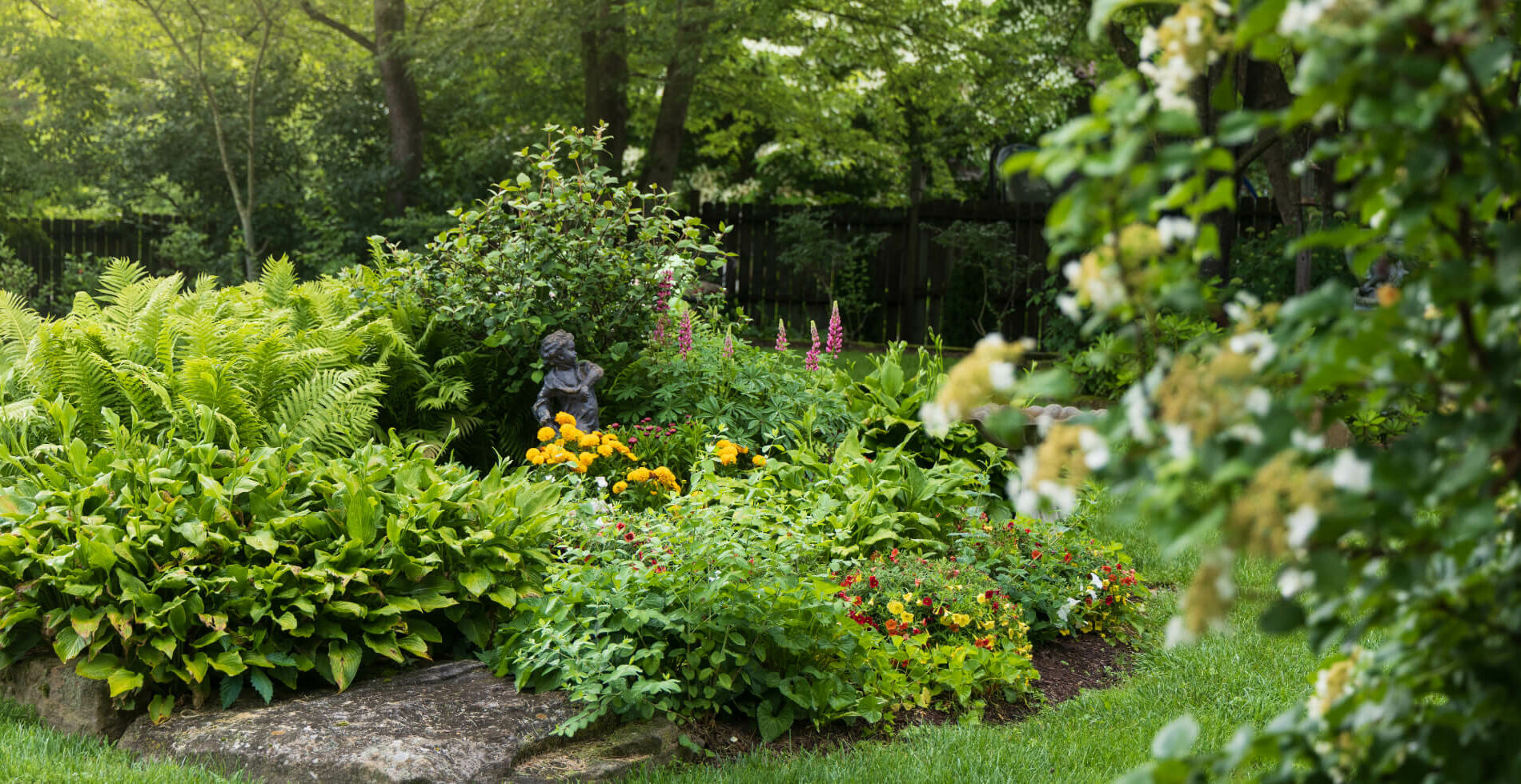 A lush garden area with mature trees and bushes, multi colored flowers, and a cherub angel statue.
