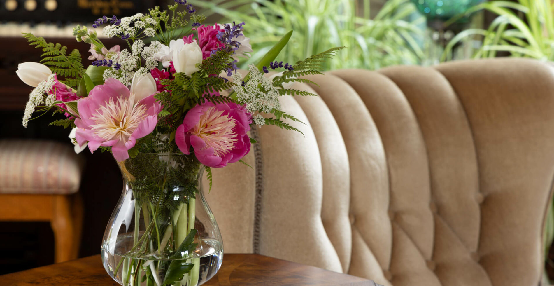 A clear vase with a bouquet of colorful flowers and greenery on a small table next to a plush tan armchair.