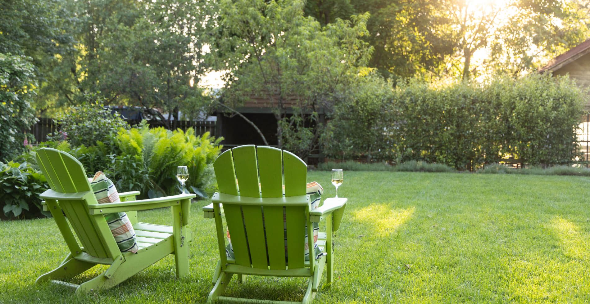 Two green adirondack chairs with glasses of white wine on the arms, set on a large grassy area looking over the lush gardens, bushes and trees.
